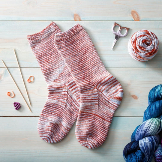 A pair of socks knit in Manos Del Uraguay Alegria, colorway Colorado River, laid flat on a wooden surface. Scissors, knitting notions and yarn surround the socks. 