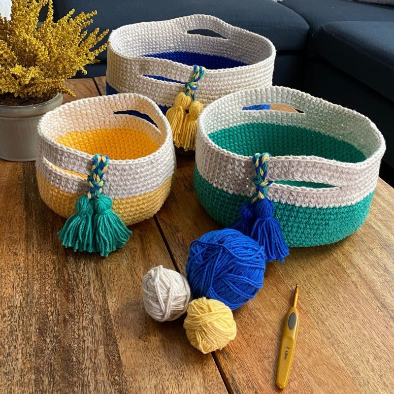 A set of three baskets crocheted from Cascade Yarns Nifty Cotton in colors Blue, Yellow, Green and Natural are shown, along with a few balls of yarn and a crochet hook.