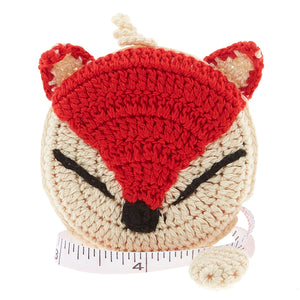 A crochet covered tape measure in the shape of a fox head with a red-orange and white face, with black nose and eyes.