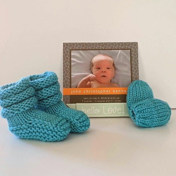 A hat, two mitts and two booties hand-knit for babies, shown with a birth announcement.
