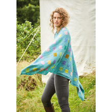 Load image into Gallery viewer, A woman in black jeans and knee high books has a turquoise knit blanket  draped over her shoulders. The blanket features an evenly spaced grid pattern of small squares in saturated pastel tones..
