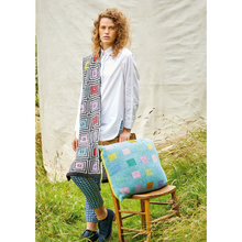 Load image into Gallery viewer, A woman stands in long grass before a white backdrop leaning on a small wooden chair.  She is wearing a white buttondown shirt and a knit scarf over one shoulder that features a bold black and white geometric pattern with pops of bright color, a knit cushion leans on the back of the chair and features evenly spaced squares in pale pastels on a turquoise background.
