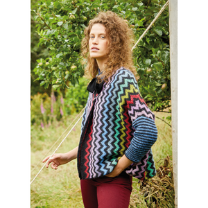 A woman in red jeans stands before an apple tree wearing a knit jacket featuring side-to-side zig zags alternating between black and bold primary and pastel colors, with simple blue and black striped sleeves.