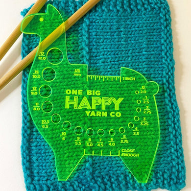 A bright yellow acrylic llama needle & hook gauge tool rests atop a turquoise knitted swatch and bamboo knitting needles. 