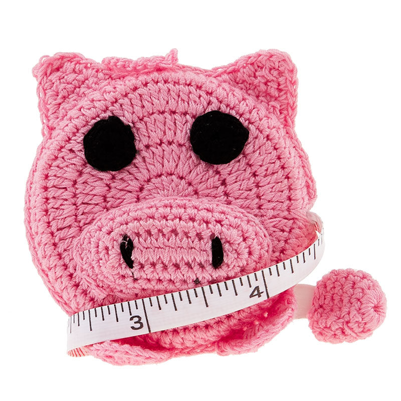 A crochet covered tape measure in the shape of a pig's face, featuring a pink face and black features.